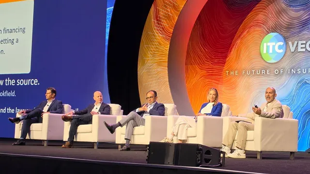 Five panelists seated in white armchairs on the Main Stage, one speaking with a slide remote, beside a screen showing a disclosure example.