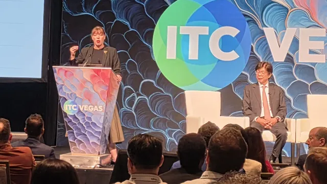 A speaker at the ITC Vegas podium while a second presenter listens from a white chair on stage.