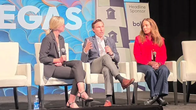A moderator and two panelists seated on white chairs on the ITC Vegas stage, the center panelist speaking into a microphone.